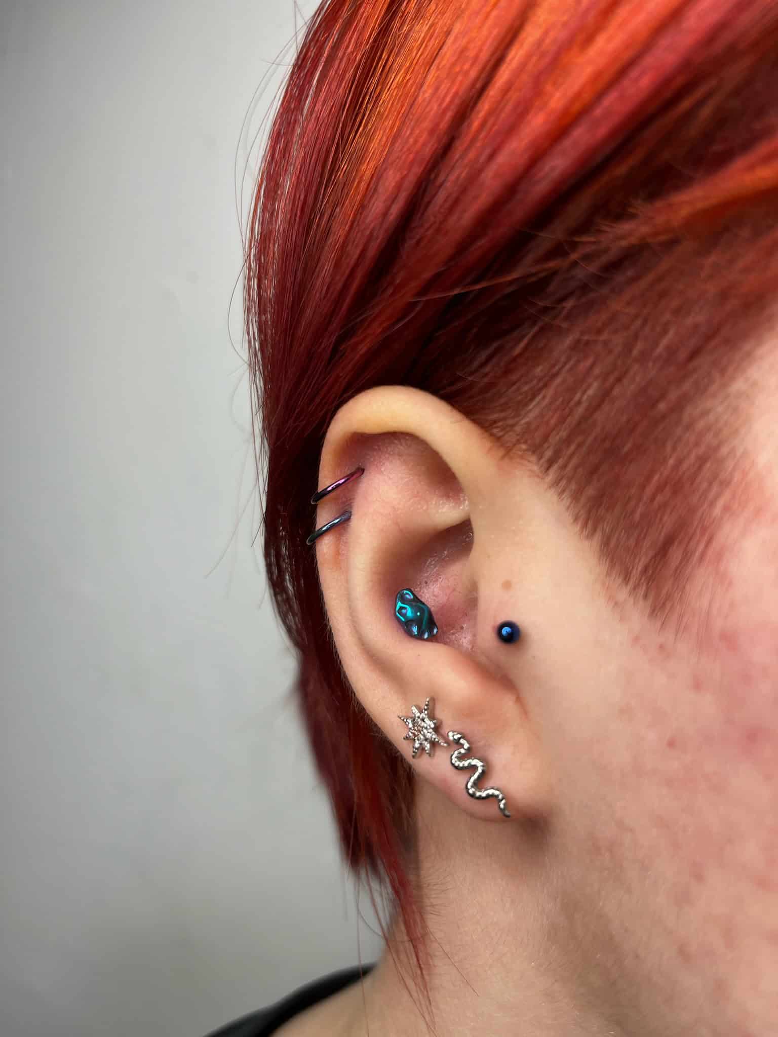 7sinstattoo.live Zbliżenie ucha osoby z wieloma piercingami i jaskrawoczerwonymi włosami. Ucho ma różnorodną biżuterię, w tym niebieski kolczyk w tragusie, mały niebieski kółko w helisie, srebrny kolczyk w kształcie węża i kolczyk w kształcie gwiazdy na płatku ucha. Odkryj więcej wyjątkowych stylów w Studio Tatuażu Poznań z IMG_6681. Najlepsze studio tatuażu Seven Sins Tattoo