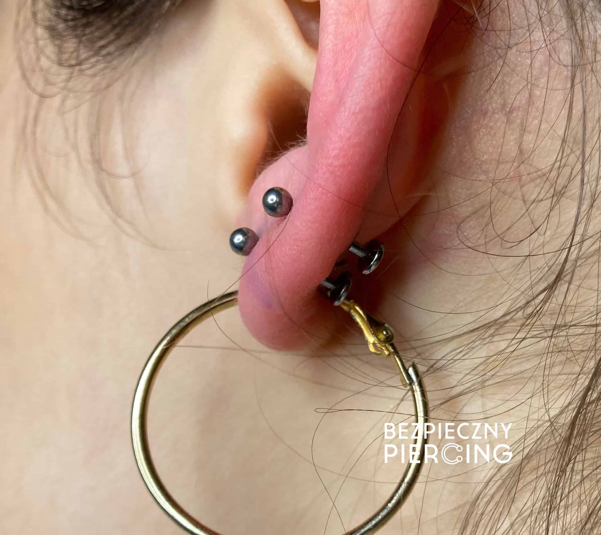 7sinstattoo.live Zbliżenie ucha osoby ozdobionego dużym piercingiem i dwoma mniejszymi piercingami nad nim, wszystkie prezentujące stylową biżuterię. Wyraźny kolczyk-kółko jest osadzony w dużym piercingu. Tekst na zdjęciu, „Bezpieczny Piercing”, podkreśla studio tatuaży Poznań jako najlepszy wybór w zakresie modnej sztuki ciała. Produkt: IMG_6695 Najlepsze studio tatuażu Seven Sins Tattoo