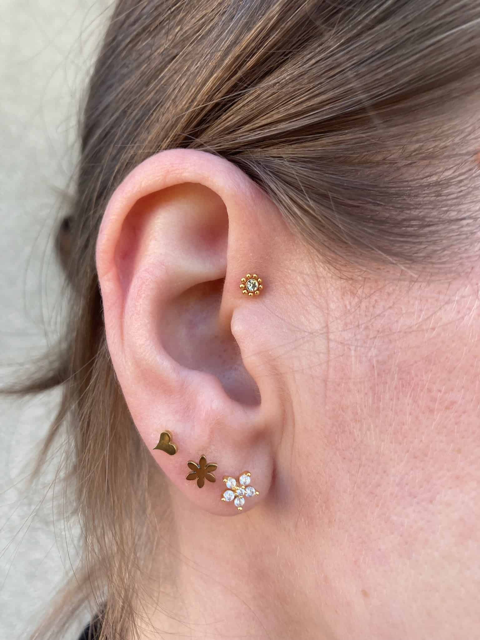 7sinstattoo.live Zbliżenie ucha osoby ozdobionego wieloma piercingami, przedstawiające produkt IMG_6892. Górna chrząstka ucha prezentuje mały kolczyk w kształcie kwiatka, podczas gdy dolny płatek ucha prezentuje trzy kolczyki: małe serce, gwiazdę i kwiat ozdobiony kamieniami szlachetnymi. Brązowe włosy osoby są schowane za uchem, tworząc artystyczny wygląd przypominający studio tatuaży Poznań. Najlepsze studio tatuażu Seven Sins Tattoo