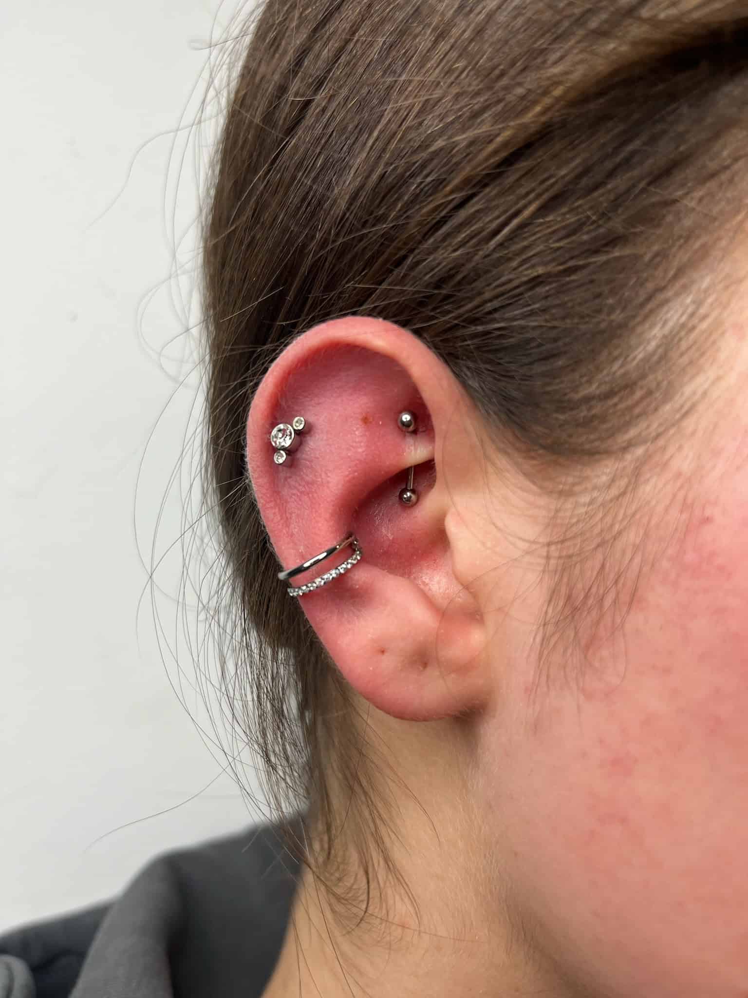 7sinstattoo.live Zbliżenie ucha z wieloma piercingami ze studia tatuaży Poznań (IMG_7134). W chrząstce znajdują się dwa kolczyki-sztyfty, a w płatku dwa kolczyki-kółka, z których jeden ma rząd małych kamieni. Ucho ma delikatne zaczerwienienie wokół piercingu, widoczne są brązowe włosy i jasna skóra. Najlepsze studio tatuażu Seven Sins Tattoo
