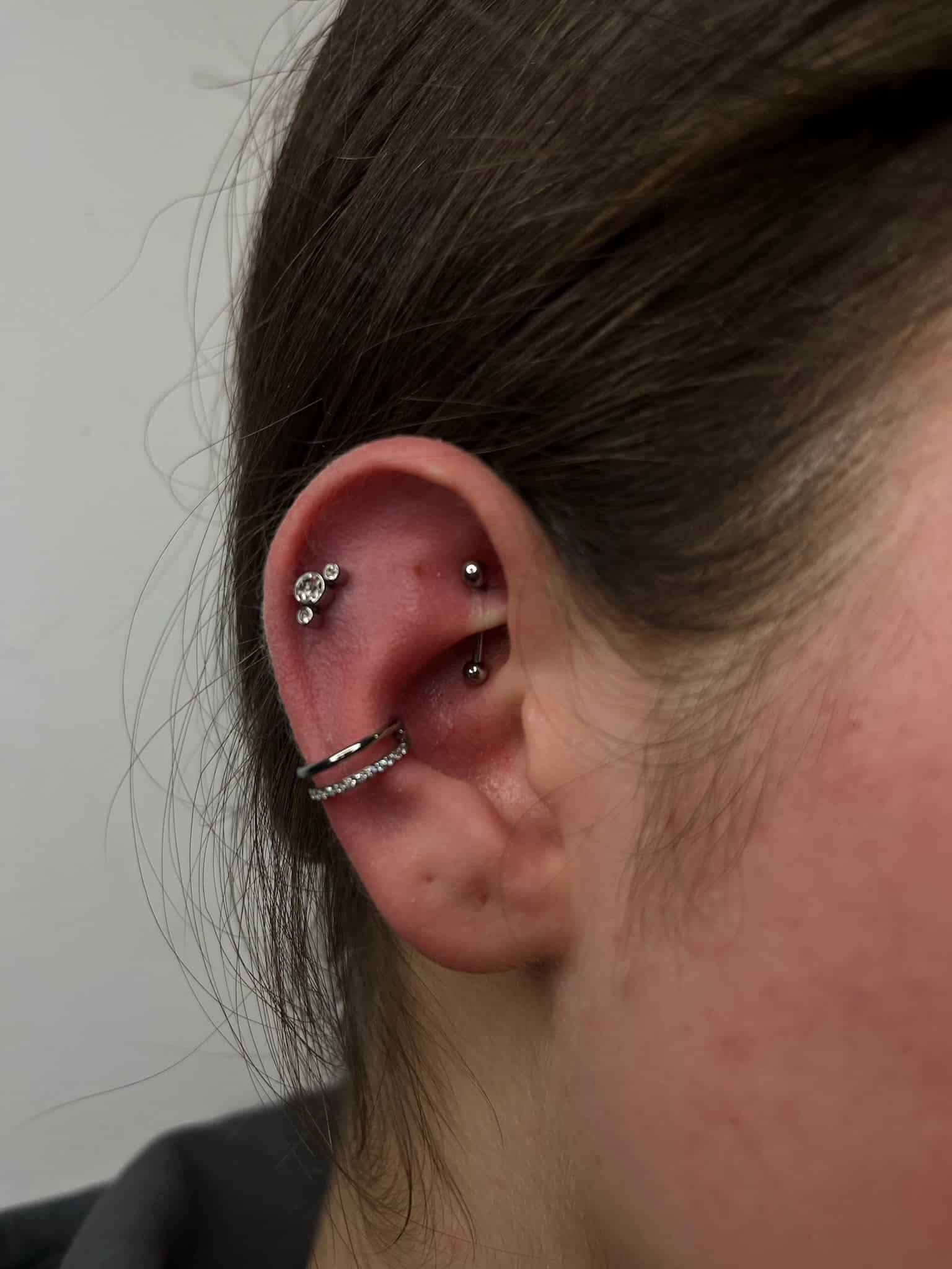 7sinstattoo.live Zbliżenie na ucho osoby z wieloma piercingami, wykonane w najlepszym studio tatuażu w Poznaniu, z IMG_7136. Piercingi obejmują mały kolczyk z wzorem w kształcie kwiatu na górnej części ucha, zakrzywiony pręt w chrząstce i kolczyk-kółko z osadzonymi klejnotami w płatku ucha. Ucho jest osadzone na jednolitym tle. Najlepsze studio tatuażu Seven Sins Tattoo