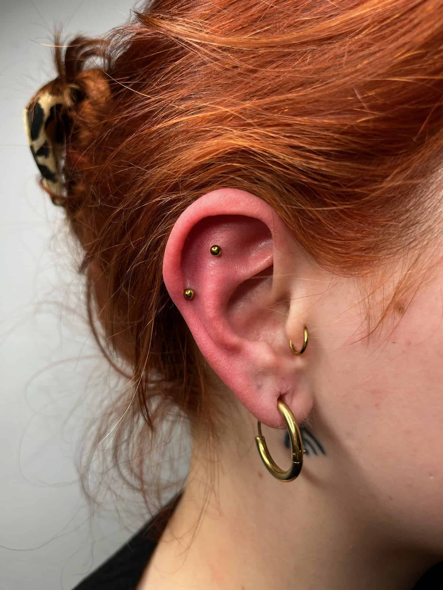 7sinstattoo.live Zbliżenie ucha osoby z wieloma piercingami, prezentujące IMG_7293, w tym złoty kółko w płatku ucha, mniejszy kółko w górnym uchu i dwa złote kolczyki na wewnętrznej chrząstce. Osoba ma rude włosy związane brązową, wzorzystą gumką do włosów. Po inspirację do sztuki na ciele odwiedź Studio Tatuaże Poznań. Najlepsze studio tatuażu Seven Sins Tattoo