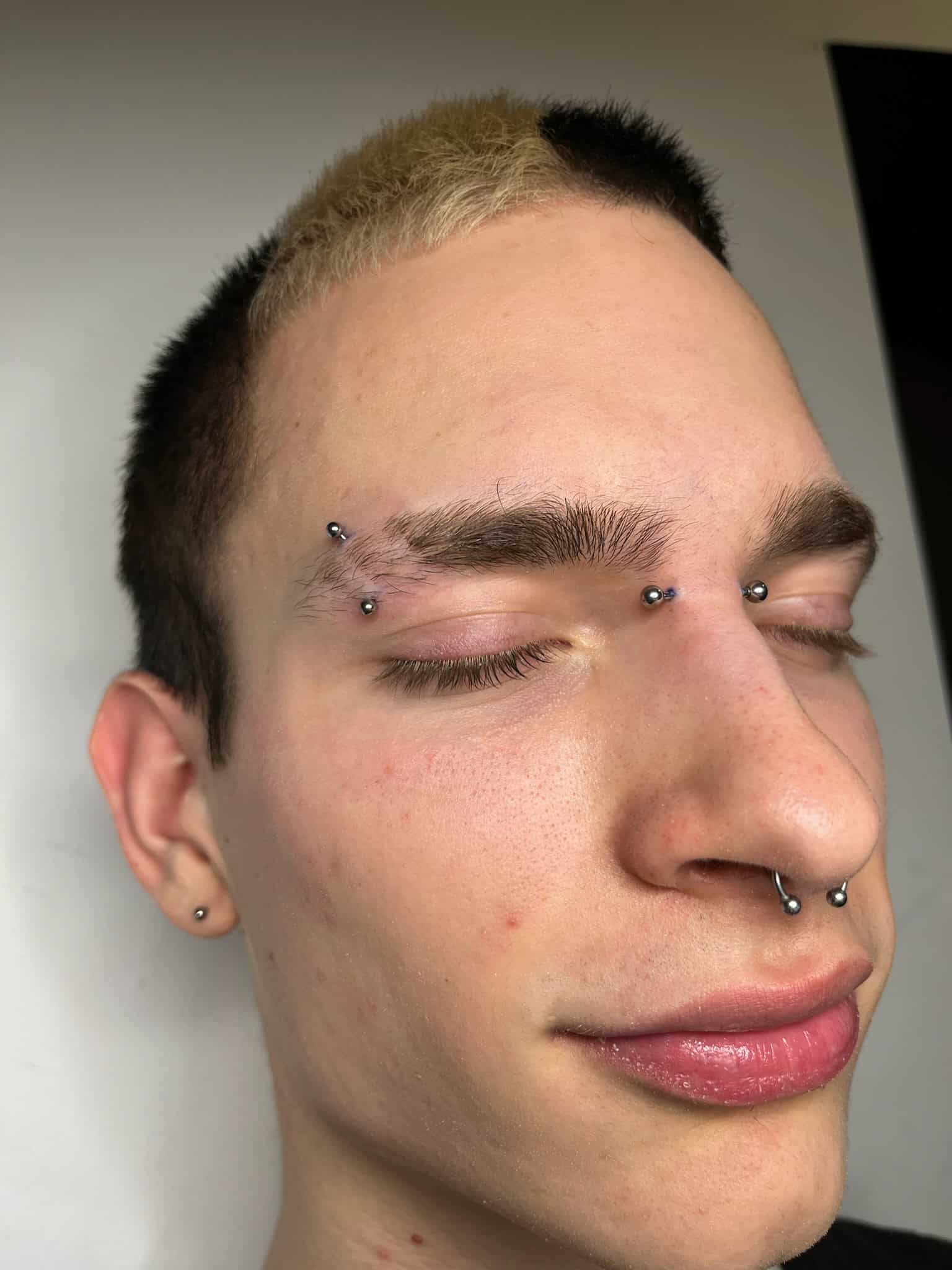 7sinstattoo.live Na zdjęciu zatytułowanym IMG_7310 osoba z krótkimi, dwukolorowymi włosami (brązowymi i rozjaśnionymi blond) jest przedstawiona z zamkniętymi oczami. Ma wiele kolczyków na twarzy: dwa na brwi, jeden między brwiami i piercing przegrody nosowej. Jej wyraz twarzy wydaje się zadowolony lub spokojny wewnątrz studio tatuażu Poznań. Najlepsze studio tatuażu Seven Sins Tattoo