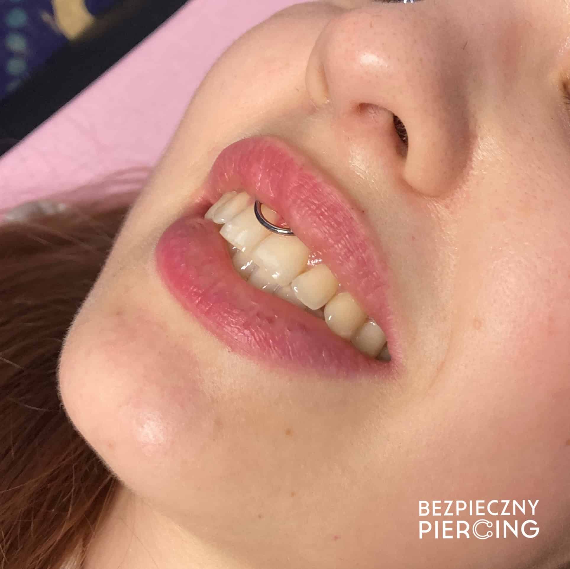 7sinstattoo.live Zbliżenie twarzy osoby pochylonej ku górze, ukazującej uśmiech ozdobiony srebrnym kółkiem w wardze i jasnoróżową szminką, z napisem „BEZPIECZNY PIERCING” w prawym dolnym rogu, ilustrującym wysokie standardy utrzymywane w studio tatuażu Poznań. Obraz nosi tytuł IMG_7516. Najlepsze studio tatuażu Seven Sins Tattoo