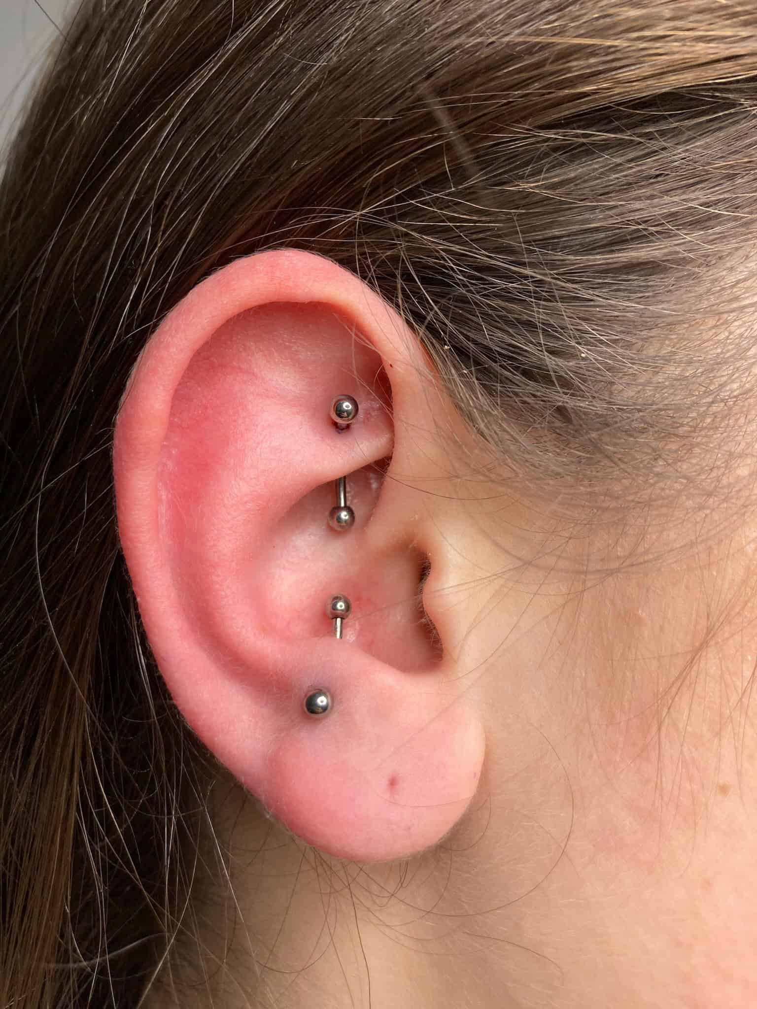7sinstattoo.live Zbliżenie na ucho osoby z wieloma piercingami, podkreślające kunszt najlepszego studia tatuażu Poznań. Są trzy srebrne ćwieki: jeden w helisie (górna część), jeden w tragusie (blisko kanału słuchowego) i jeden w płacie ucha. Ucho wygląda zdrowo, bez oznak podrażnienia lub zaczerwienienia wokół piercingu, idealnie zamknięte w IMG_7546. Najlepsze studio tatuażu Seven Sins Tattoo
