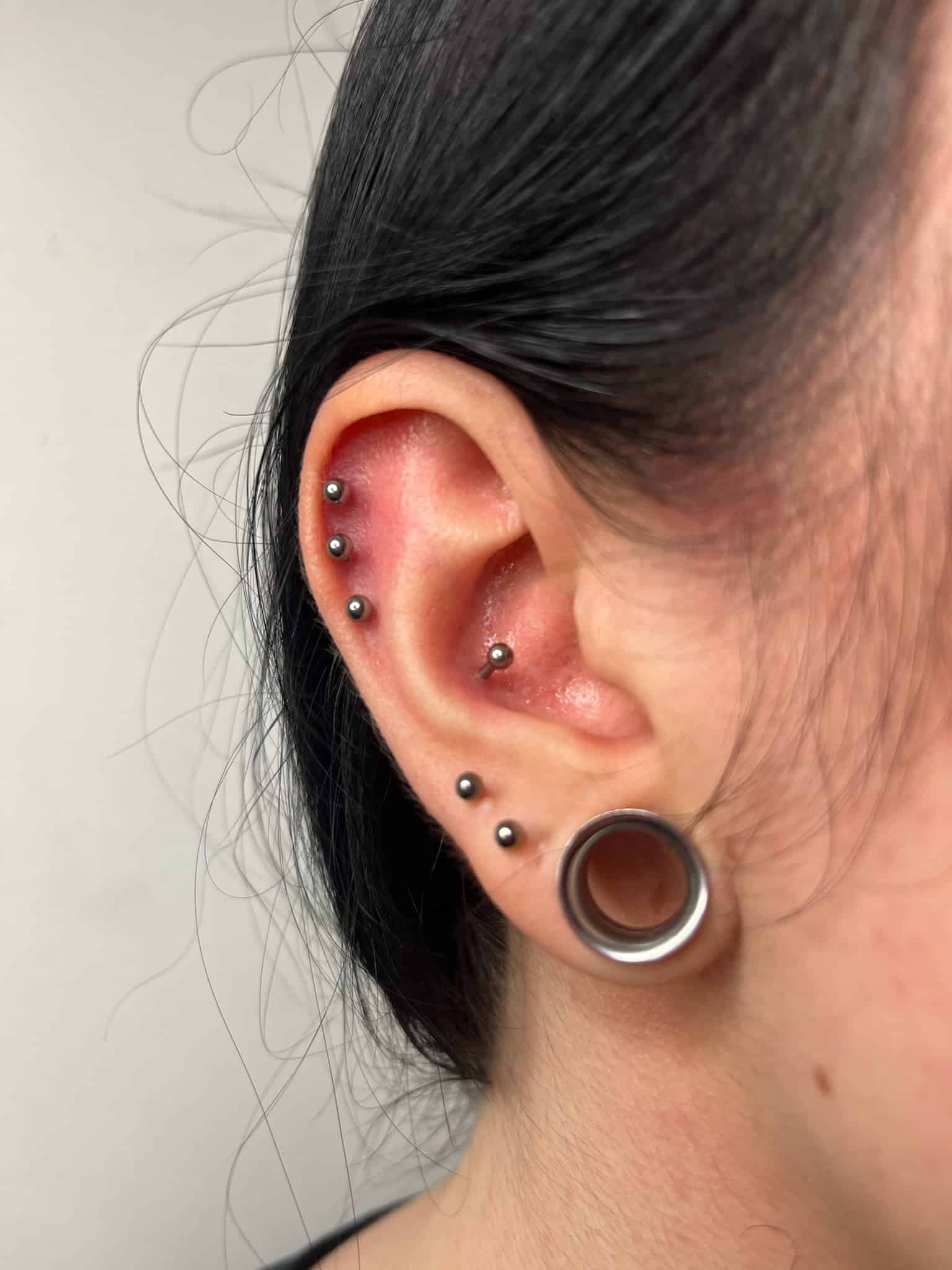 7sinstattoo.live Zbliżenie ucha osoby z wieloma piercingami uchwyconymi w IMG_7709 w Studio Tatuaże Poznań. Ucho ma srebrne ćwieki zdobiące zarówno płatek ucha, jak i helisę, a także duży korek w płatku ucha. Skóra wokół piercingu wydaje się lekko czerwona, a osoba ma ciemne włosy. Najlepsze studio tatuażu Seven Sins Tattoo