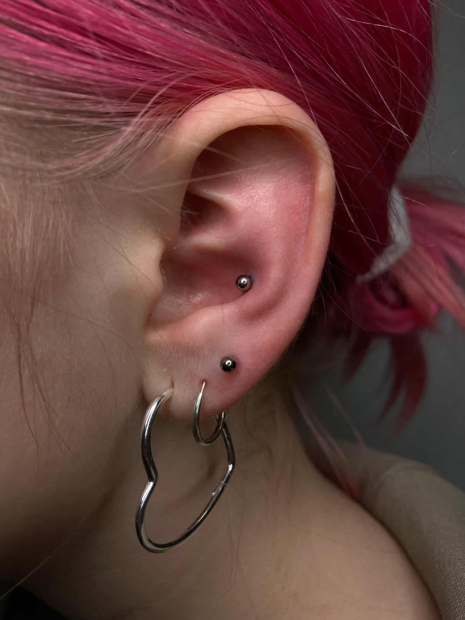 7sinstattoo.live Zbliżenie ucha osoby z jasnoróżowymi włosami, z dwoma małymi czarnymi kolczykami w chrząstce i srebrnym kolczykiem w płatku ucha, uchwyconym w IMG_7737. Tło jest neutralne i nieostre, zapewniając idealne tło do zaprezentowania nowego tuszu ze studia Tatuaże Poznań. Najlepsze studio tatuażu Seven Sins Tattoo