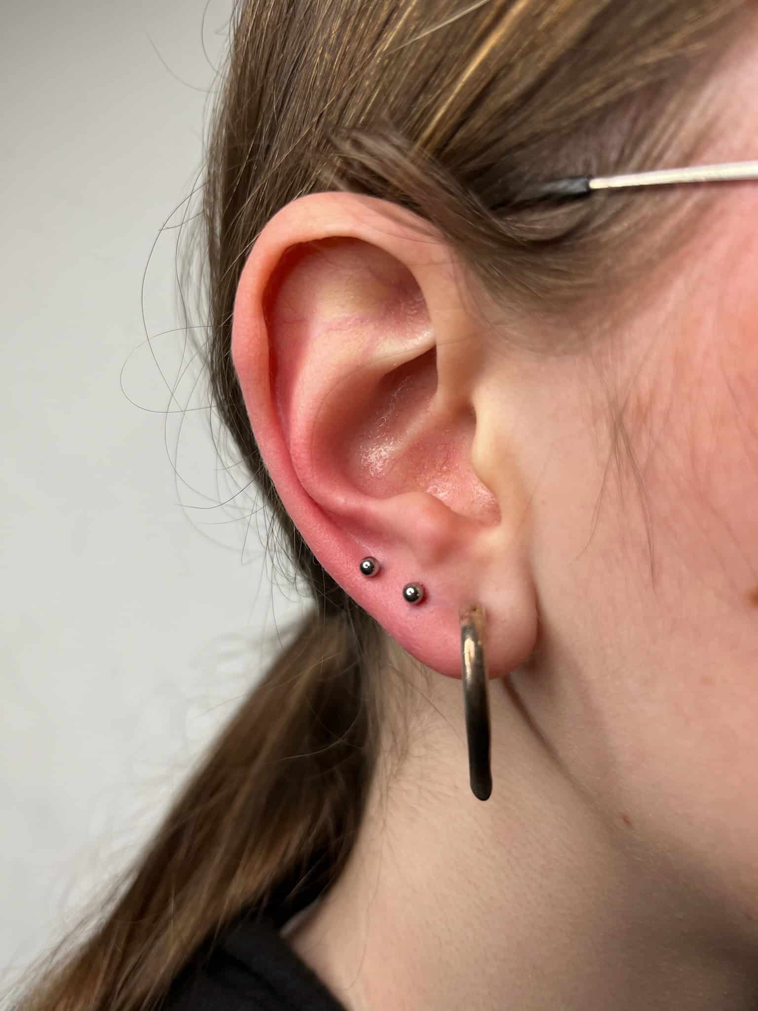 7sinstattoo.live Zbliżenie ucha osoby z trzema kolczykami: dwoma małymi ćwiekami w górnym płacie i jednym dużym kółkiem w dolnym płacie. Osoba ta, prawdopodobnie odwiedzająca studio tatuażu w Poznaniu, ma brązowe włosy związane z tyłu i widoczną oprawkę okularów. Plik obrazu oznaczony jako IMG_7744. Najlepsze studio tatuażu Seven Sins Tattoo