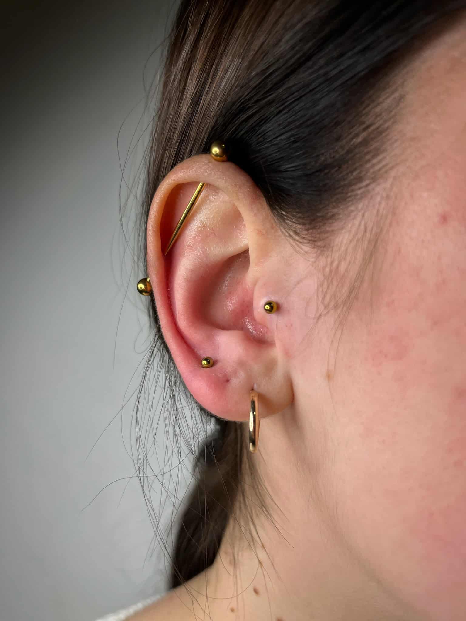 7sinstattoo.live Zbliżenie na ucho osoby z pięcioma przekłuciami, z różną złotą biżuterią z IMG_8481. Przekłucia obejmują standardowy piercing płatka ucha, przekłucie helix z kółkiem oraz dwa dodatkowe przekłucia helix i przekłucie industrialne, wszystkie ozdobione małymi złotymi ćwiekami i sztabkami z najlepszego studia tatuażu Poznań. Najlepsze studio tatuażu Seven Sins Tattoo