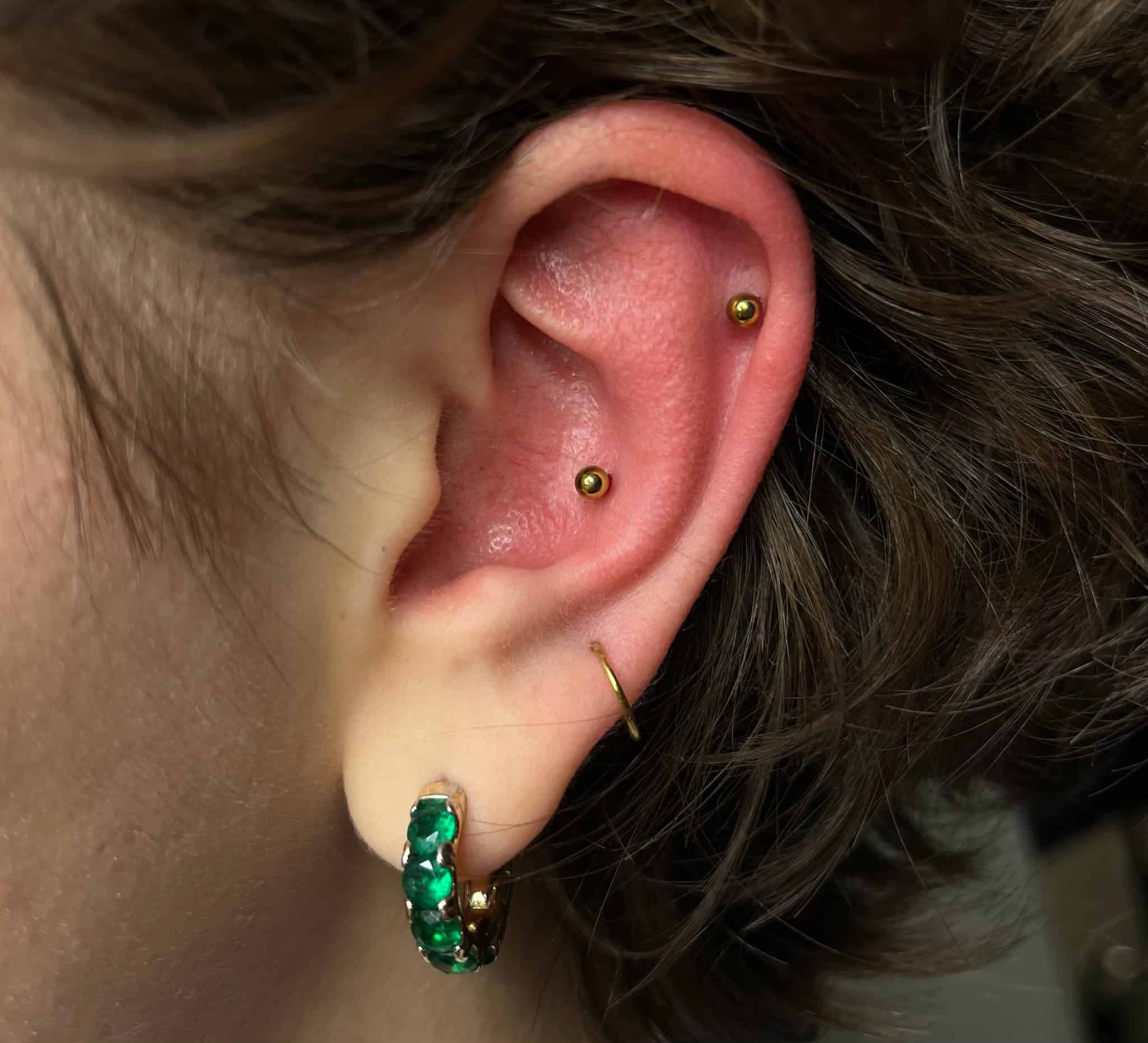 7sinstattoo.live Zbliżenie ucha osoby ozdobionego wieloma piercingami, zrobione w jednym z najlepszych studiów tatuażu w Poznaniu. Ucho ma złoty kolczyk w okolicy muszli, złoty kolczyk w górnym płatku ucha, mały złoty pierścień w płatku ucha i zielony koralikowy kolczyk w kształcie kółka w dolnym płatku ucha. Obraz nosi tytuł IMG_8588. Najlepsze studio tatuażu Seven Sins Tattoo