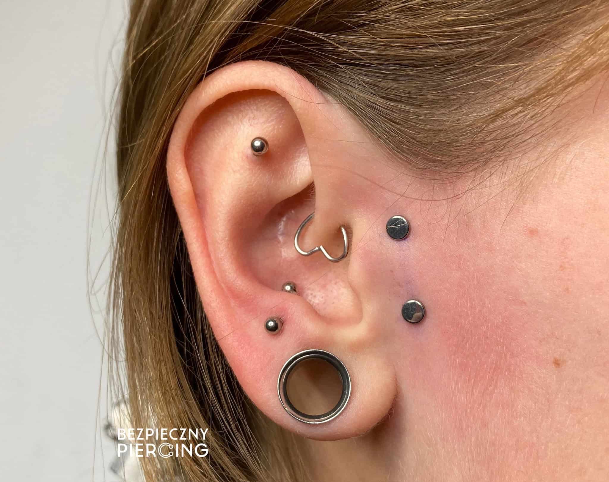 7sinstattoo.live Zbliżenie ucha osoby z wieloma piercingami, w tym kółkiem w przedniej helisie, ćwiekiem w górnej helisie, kółkiem w muszli, ćwiekiem w tragusie i dwoma ćwiekami w płatku ucha wraz z tunelem. Tekst „BEZPIECZNY PIERCING” znajduje się w lewym dolnym rogu. Odwiedź nasze studio tat (Nazwa produktu: IMG_8634). Najlepsze studio tatuażu Seven Sins Tattoo