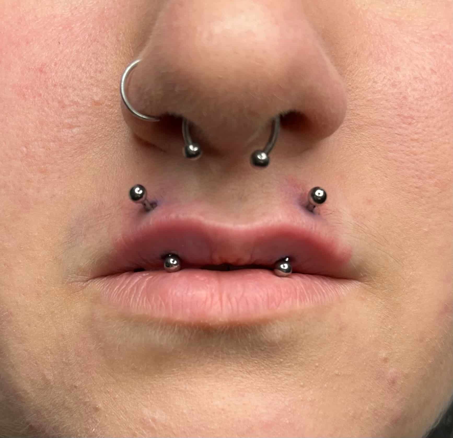 7sinstattoo.live Zbliżenie ust i nosa osoby, prezentujące kilka piercingów. Osoba ma dwa piercingi po każdej stronie dolnej wargi i dwa w środku, a także piercing przegrody nosowej i kolczyk w nozdrzu po lewej stronie. Ten uderzający wygląd został dopracowany w studio tatuaży Poznań, jak uchwycono w produkcie IMG_8928. Najlepsze studio tatuażu Seven Sins Tattoo