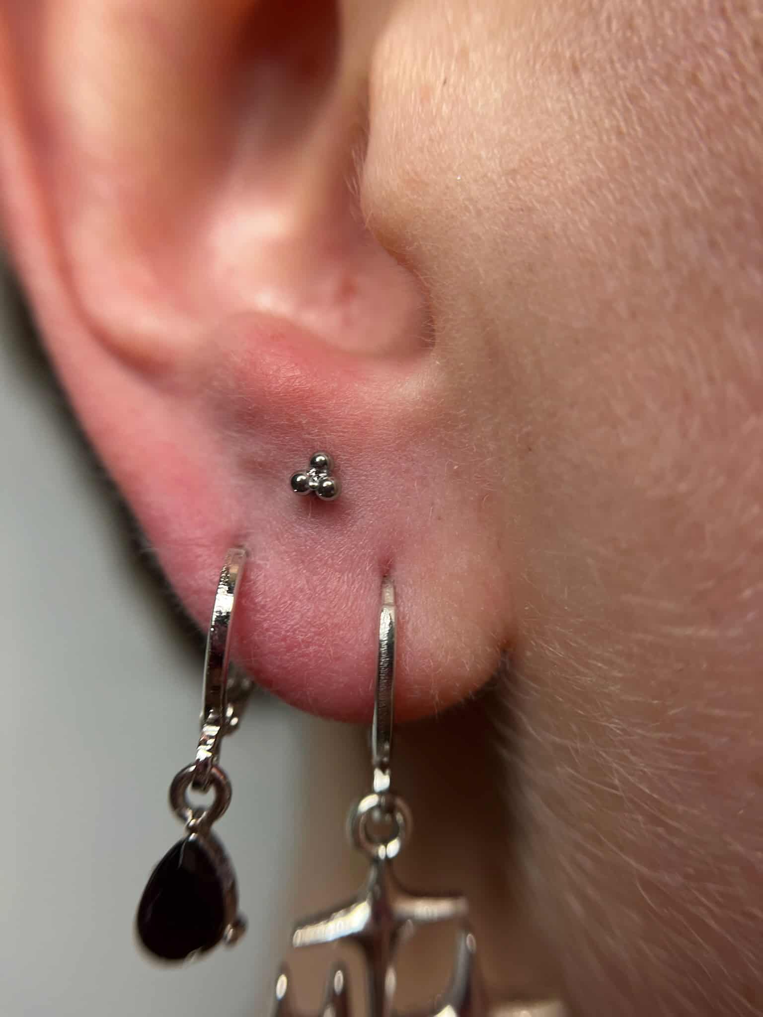 7sinstattoo.live Zbliżenie na ucho z dwoma piercingami, ozdobione w studio tatuażu Poznań. Jeden piercing ma mały srebrny ćwiek z trzema malutkimi kulkami, podczas gdy drugi prezentuje IMG_9004, srebrny kolczyk-kółko z czarnym łezkowatym urokiem i zwisającym urokiem wagi. Najlepsze studio tatuażu Seven Sins Tattoo