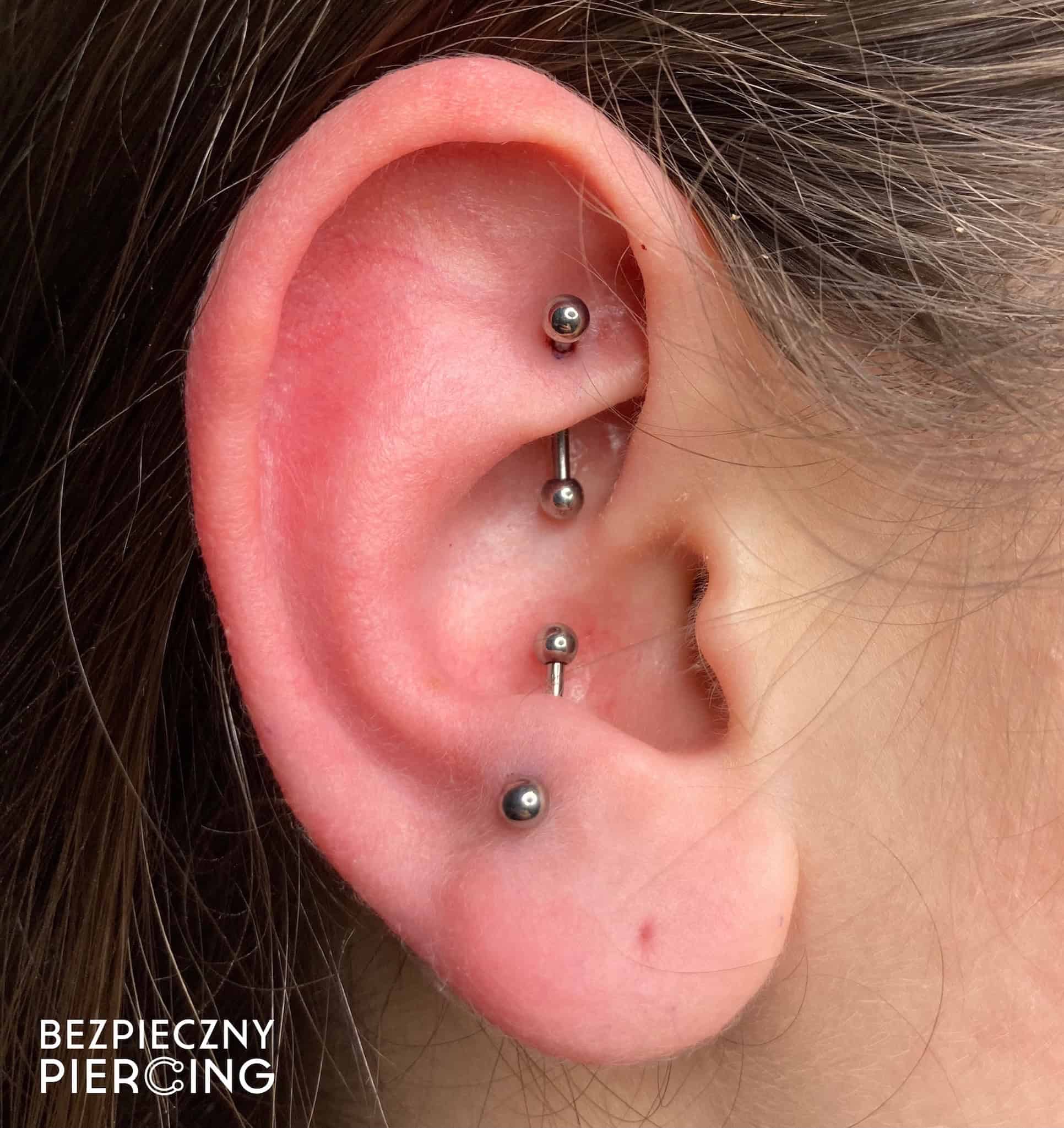 7sinstattoo.live Zbliżenie na ucho osoby z wieloma piercingami, w tym trzema srebrnymi ćwiekami w chrząstce i jednym w płatku ucha. Tekst w lewym dolnym rogu brzmi „BEZPIECZNY PIERCING”. Skóra wokół piercingu wygląda na zdrową i dobrze zagojoną, co świadczy o doświadczeniu najlepszego studia tatuażu w Poznaniu. Obraz oznaczony jako IMG_9083. Najlepsze studio tatuażu Seven Sins Tattoo