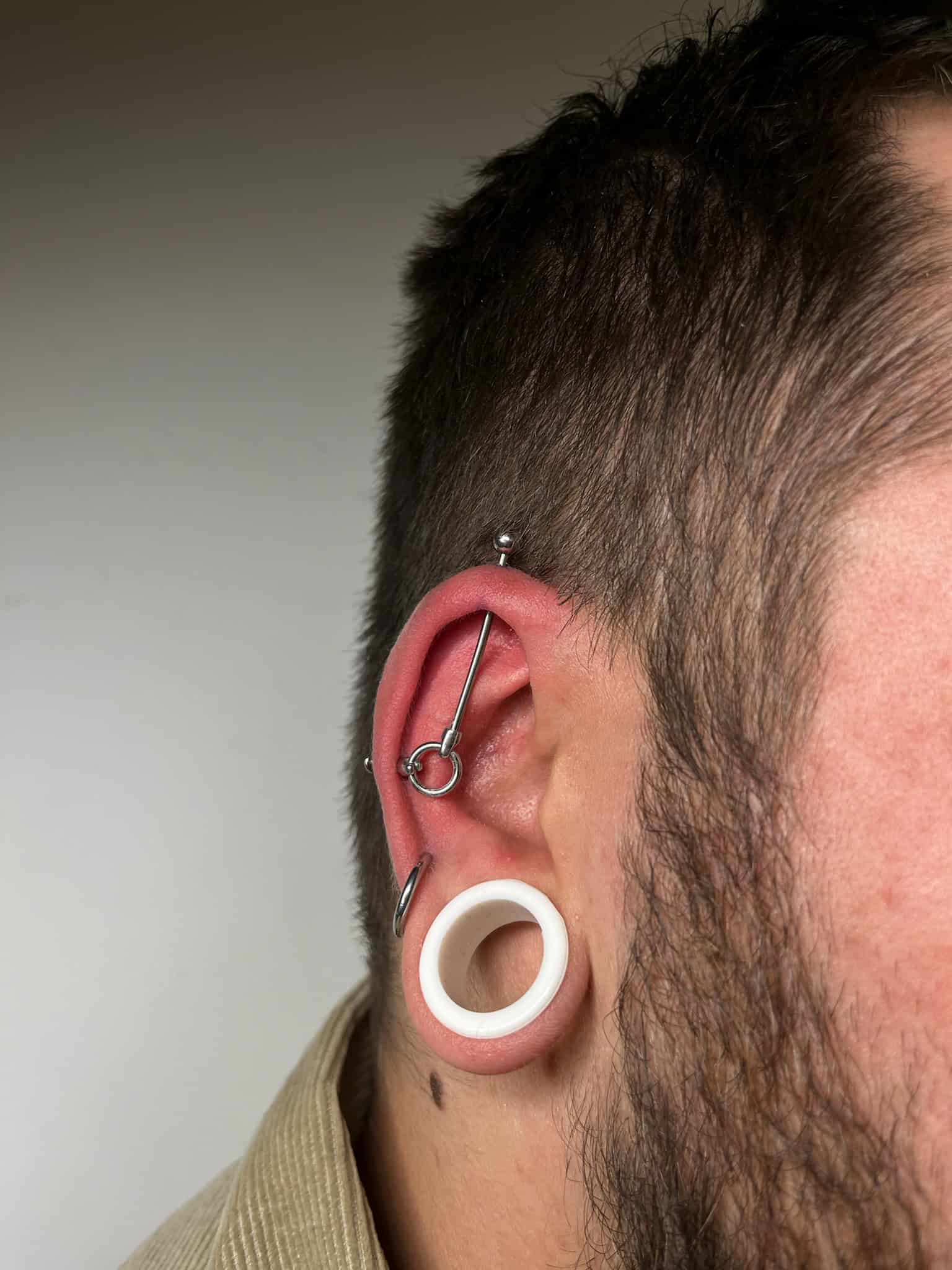 7sinstattoo.live Zbliżenie ucha osoby ozdobionego wieloma piercingami, idealne dla każdego odwiedzającego najlepsze studio tatuażu w Poznaniu. Ucho ma duży biały wskaźnik w płatku ucha, kółko w helisie, srebrne kółko z małą kulką w muszli i kolczyk sztangi w górnej chrząstce ucha. Tło jest gładkie. Nazwa produktu: IMG_9597. Najlepsze studio tatuażu Seven Sins Tattoo