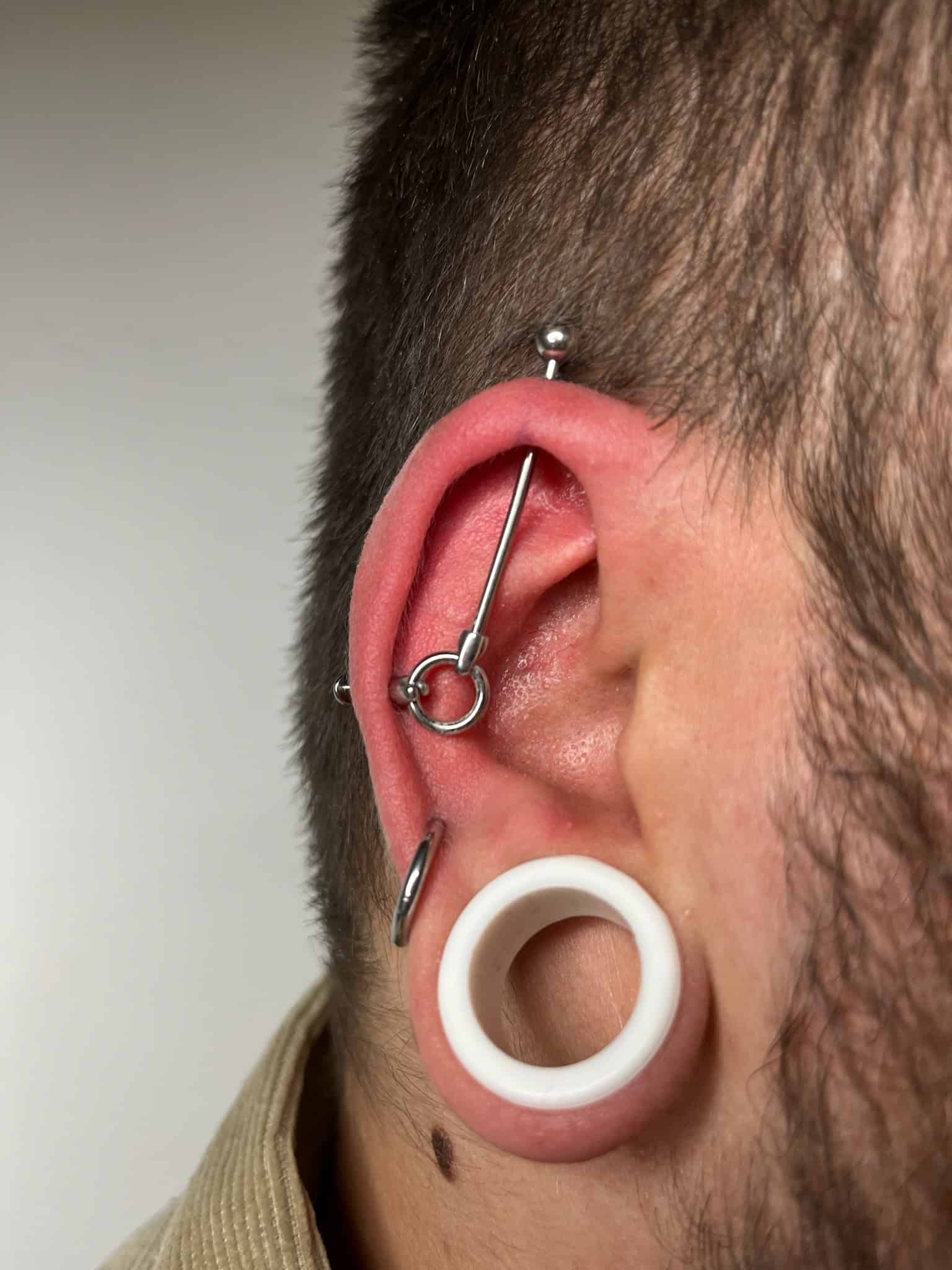 7sinstattoo.live Zbliżenie ucha osoby pokazuje kilka piercingów: biały okrągły wskaźnik w płatku ucha, sztangę w górnej chrząstce ucha i pierścień w muszli. Ucho wydaje się lekko czerwone, a osoba ma krótkie włosy. Ta niedawna praca, przedstawiona w produkcie IMG_9599, została wykonana w najlepsze studio tatuażu Poznań. Najlepsze studio tatuażu Seven Sins Tattoo
