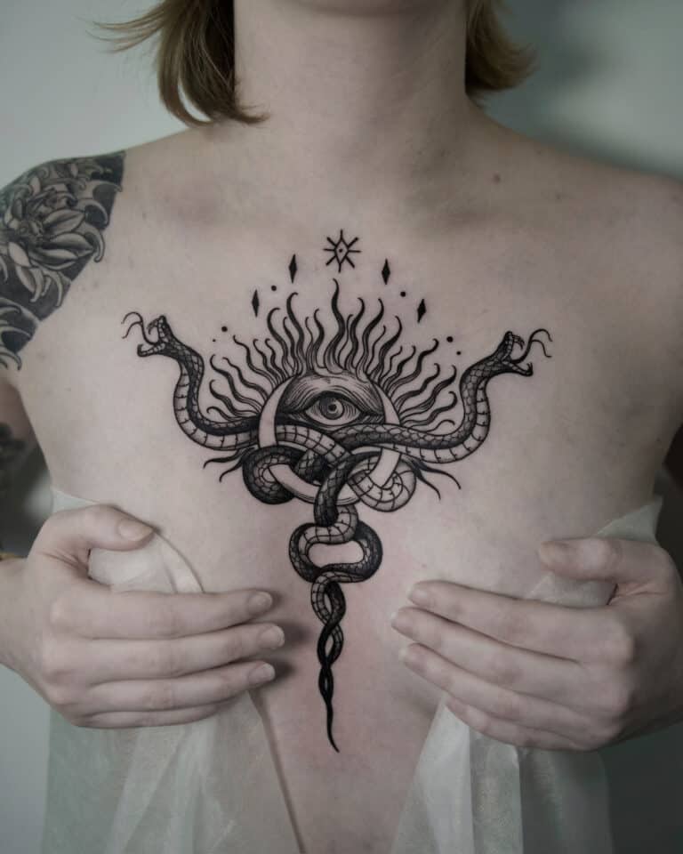7sinstattoo.live Osoba z dużym tatuażem na klatce piersiowej przedstawiającym oko otoczone płomieniami i splecionymi wężami trzyma lekką, półprzezroczystą tkaninę, częściowo zakrywającą dolną część tatuażu. Czarno-szary tatuaż kwiatowy, stworzony przez studio tatuażu Poznań, zdobi jej górną część lewego ramienia. Ten element nosi nazwę jsnhj. Najlepsze studio tatuażu Seven Sins Tattoo