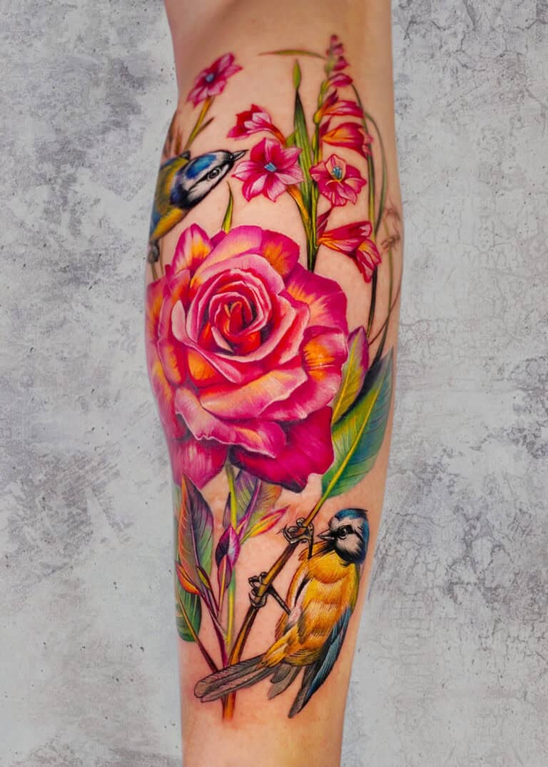 7sinstattoo.live Żywy tatuaż na ramieniu osoby, wykonany w najlepszym studio tatuażu w Poznaniu, przedstawia dużą różową różę otoczoną mniejszymi różowymi kwiatami, zielonymi liśćmi i dwoma niebieskimi i żółtymi ptakami. Szczegółowy projekt ożywa dzięki realistycznej fakturze na neutralnym, teksturowanym szarym tle. Ta oszałamiająca grafika jest znana jako produkt x39. Najlepsze studio tatuażu Seven Sins Tattoo