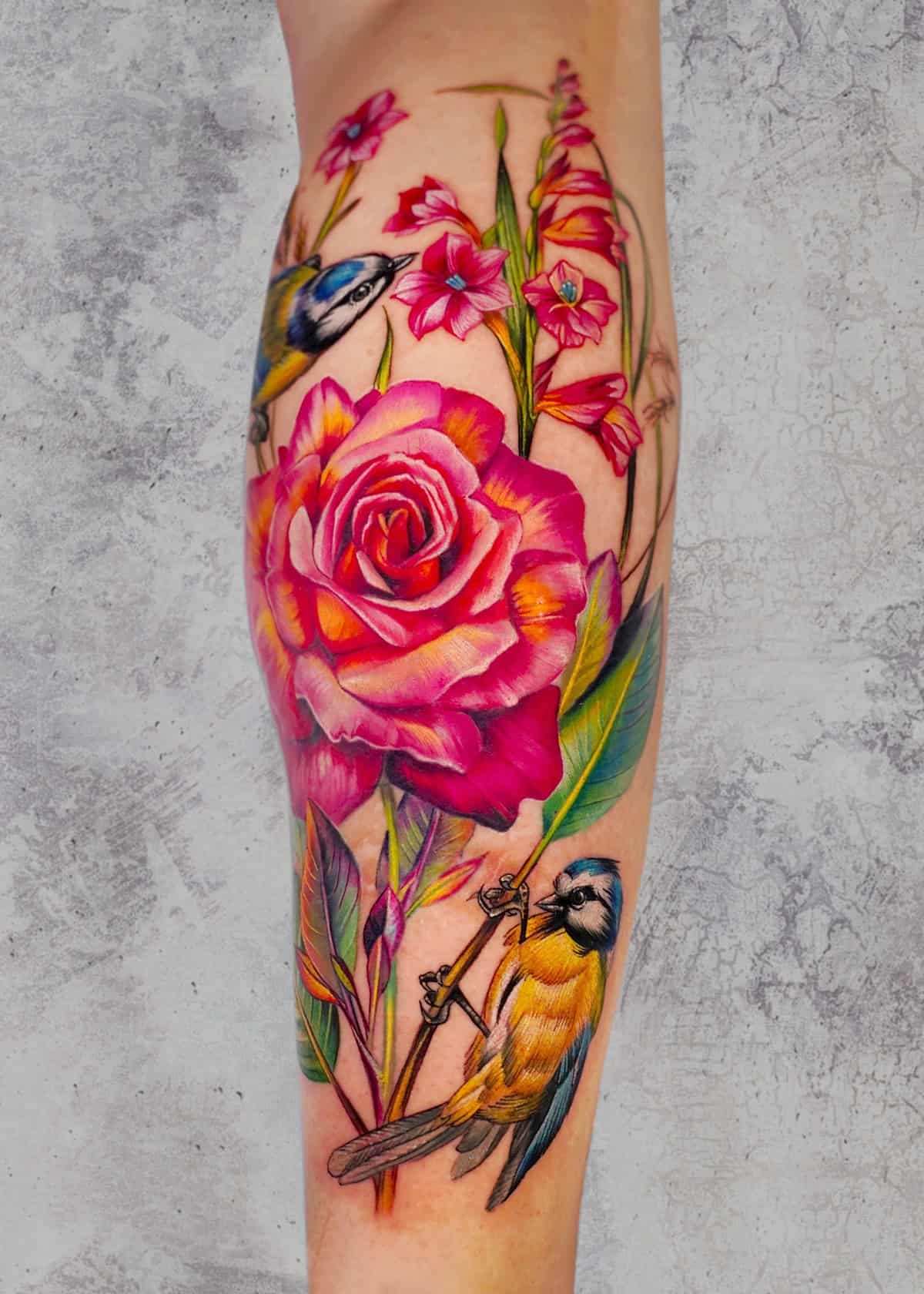 7sinstattoo.live Żywy tatuaż na ramieniu osoby, wykonany w najlepszym studio tatuażu w Poznaniu, przedstawia dużą różową różę otoczoną mniejszymi różowymi kwiatami, zielonymi liśćmi i dwoma niebieskimi i żółtymi ptakami. Szczegółowy projekt ożywa dzięki realistycznej fakturze na neutralnym, teksturowanym szarym tle. Ta oszałamiająca grafika jest znana jako produkt x39. Najlepsze studio tatuażu Seven Sins Tattoo