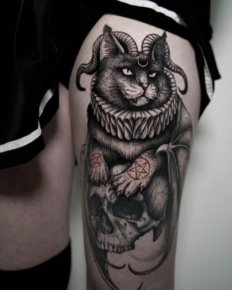 7sinstattoo.live Szczegółowy tatuaż, uchwycony na zdjęciu produktu „Копия DSC09932” przez utalentowanego tatuażystę z Poznania, przedstawia kota z rogami barana i misterną obrożą, spoczywającego na ludzkiej czaszce. Na łapach kota znajdują się dekoracyjne symbole. Tatuaż jest umieszczony na udzie osoby, z widocznym brzegiem jej czarnego ubrania. Najlepsze studio tatuażu Seven Sins Tattoo