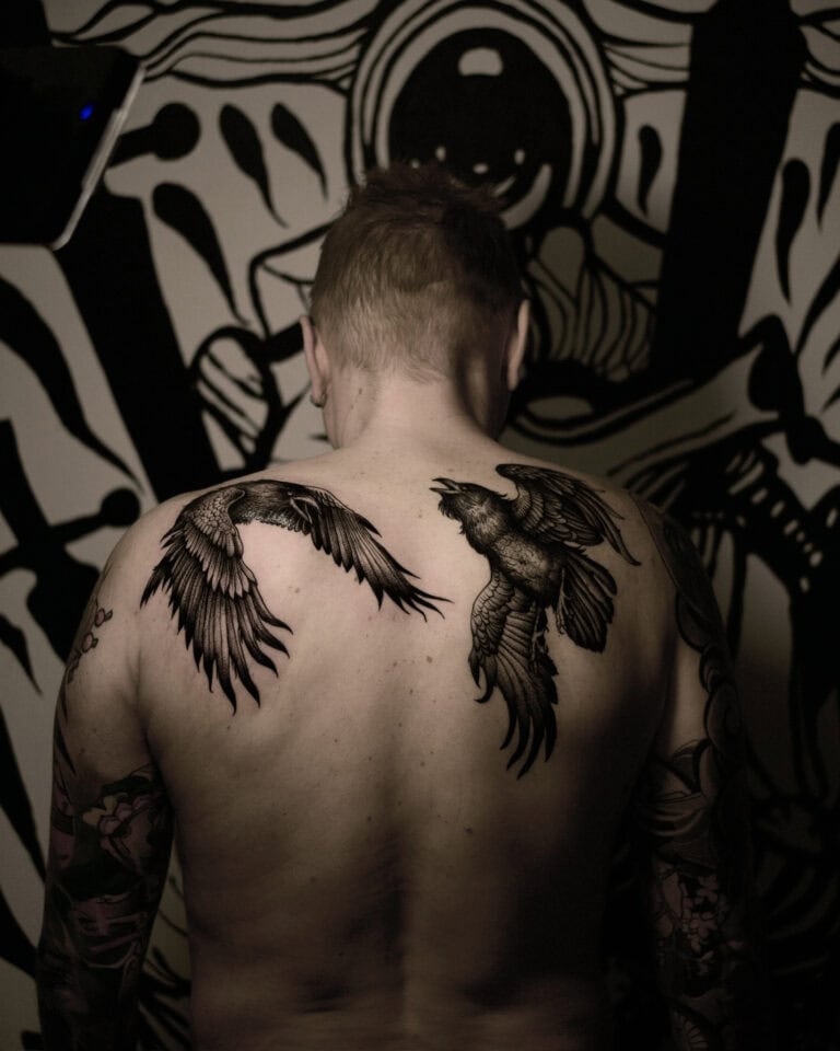 7sinstattoo.live Osoba z krótkimi włosami i widocznymi tatuażami na ramionach jest widziana od tyłu, prezentując dwa duże tatuaże ptaków na górnej części pleców. Tło przedstawia czarno-białą abstrakcyjną sztukę muralową. Obraz ma słabe, dramatyczne oświetlenie, przedstawiające wykwintną pracę najlepszego studia tatuażu Poznań. Najlepsze studio tatuażu Seven Sins Tattoo