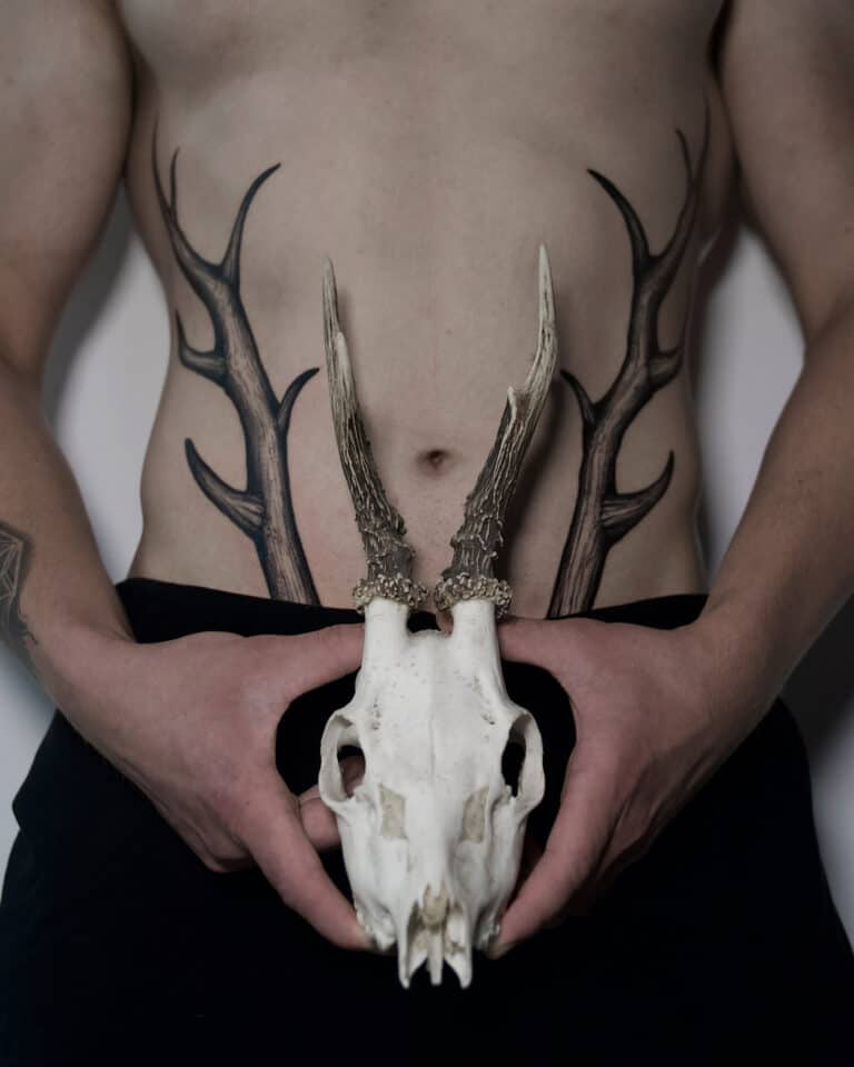7sinstattoo.live Osoba bez koszulki trzyma w rękach czaszkę jelenia, wyrównując jej poroże z misternymi czarnymi tatuażami poroża na brzuchu, tworząc iluzję, w której tatuaże płynnie rozciągają się od czaszki. Mają na sobie ciemne spodnie i stoją w renomowanym studiu tatuażu w Poznaniu. Ta artystyczna scena pięknie uosabia unikalny styl прррр. Najlepsze studio tatuażu Seven Sins Tattoo