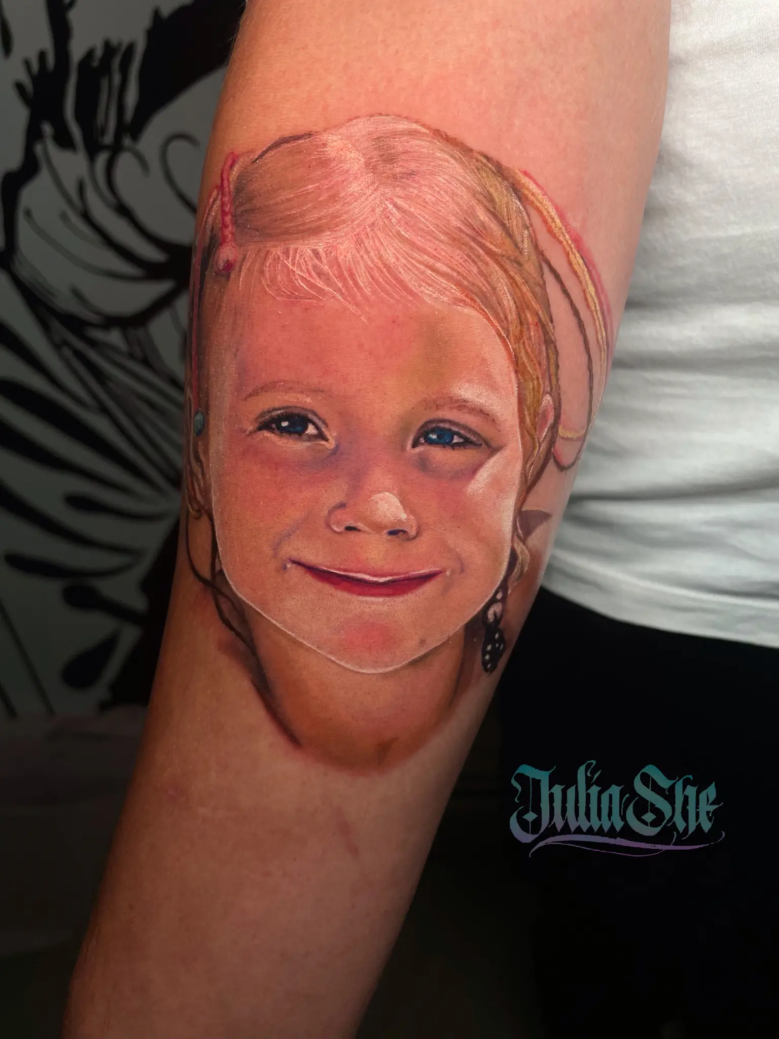 Tatuaż na ramieniu osoby przedstawia realistyczny, kolorowy portret młodej, uśmiechniętej dziewczyny o blond włosach. Tatuaż uchwyca jej rysy twarzy i wyraz w szczegółach. Pod wytatuowanym portretem imię „Julia She” jest wytatuowane stylizowaną czcionką.