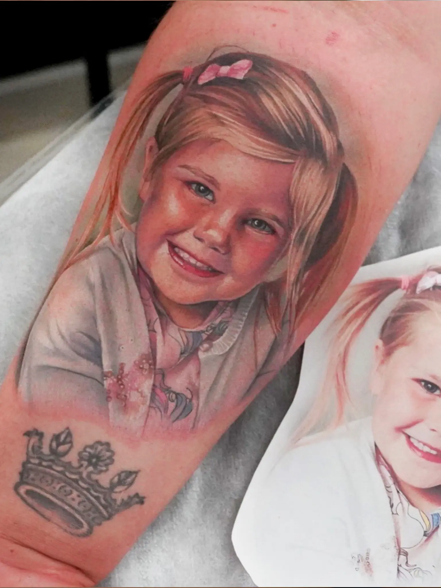Tatuaż młodej blondynki z różowymi gumkami do włosów w warkoczykach i różową kokardą zdobi ramię osoby. Obok tatuażu znajduje się papierowy wydruk oryginalnego zdjęcia dziewczyny, które tatuaż dokładnie odtwarza. Pod tatuażem znajduje się czarno-biały tatuaż korony.
