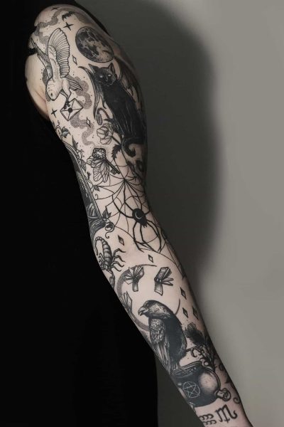 Ramię osoby zdobi misterny czarno-biały tatuaż na rękawie wykonany pieczołowicie w studiu Seven Sins Tattoo w Poznaniu. Szczegółowe wzory zawierają elementy takie jak łapacz snów, czarny kot, wrona, sowa, księżyc i różne mistyczne i geometryczne symbole na jednolitym tle. Osoba ma na sobie czarny top bez rękawów.