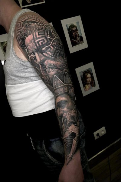 Osoba ubrana w szary podkoszulek i ciemne spodnie prezentuje misterny tatuaż na całym rękawie z motywami mitologii greckiej w Seven Sins Tattoo. Szczegółowe wzory produktu 356396244_611341101115844_5988685096994287392_n obejmują symboliczne elementy i brodatą boską postać na górze. Stoją na tle ozdobionym oprawionymi dziełami sztuki.