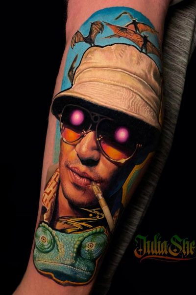Żywy tatuaż ze studia Seven Sins Tattoo w Poznaniu zdobi ramię osoby, przedstawiając osobę w kapeluszu i okularach przeciwsłonecznych, świecących różowych oczach i papierosie. Skomplikowany wzór przedstawia nietoperze latające nad nimi i kameleona poniżej, a imię „Julia She” stylowo wyryte w prawym dolnym rogu.