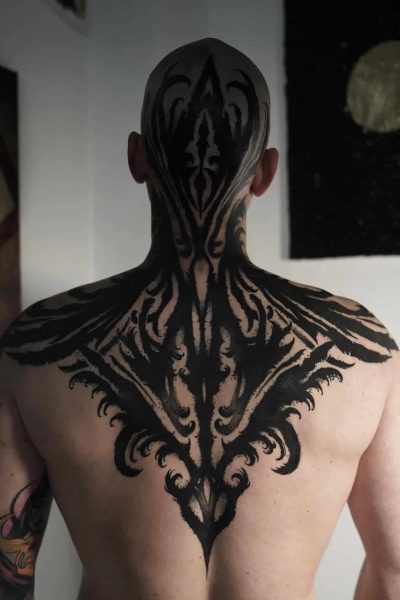 Osoba bez koszuli, z ogoloną głową, prezentuje w studiu Seven Sins Tattoo w Poznaniu misterny i czarny symy tatuaż „366065782_626882202895067_8258840146141252147_n”. Tatuaż wykonany przez utalentowanego tatuatora trzyma się od ukrytego do głowy, charakterystycznego, trzymanego wzorami z ostrymi liniami i szerokimi krzywiznami. Tło jest słabo oświetlone i ozdobione abstrakcyjną sztuką.