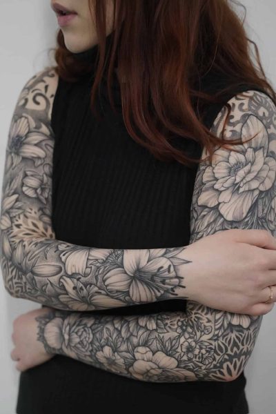 Kobieta z długimi, rudymi włosami od ramion w dół, ubrana w czarny top bez rękawów. Obie jej ręce są skrzyżowane i całkowicie pokryte szczegółowymi kwiatowymi tatuażami ze studia Seven Sins Tattoo w Poznaniu. Skomplikowane czarno-białe wzory przedstawiają różne kwiaty i liście rozciągające się od jej ramion do nadgarstków. Nosi również delikatny pierścionek na lewej ręce.