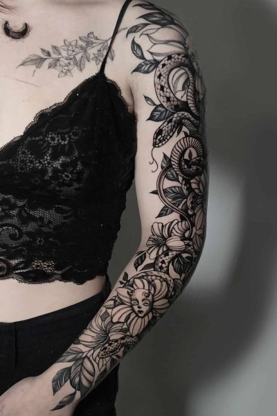 Osoba ubrana w misternie zaprojektowany czarny koronkowy top „7” i czarne spodnie z 7sinstattoo.live prezentuje szczegółowy tatuaż na lewym ramieniu. Tatuaż, wykonany w Seven Sins Tattoo Studio w Poznaniu, zawiera różne motywy kwiatowe, płynne wzory, półksiężyc na ramieniu i twarz kobiety płynnie zintegrowaną z dziełem sztuki. Dodatkowe tatuaże są również widoczne na klatce piersiowej.