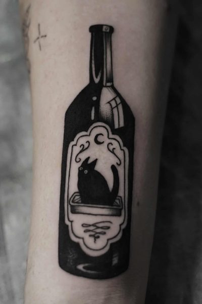 Tatuaż o nazwie DSC00008, pięknie wykonany przez utalentowanych artystów ze studia Seven Sins Tattoo w Poznaniu, przedstawia ciemną etykietę wina z sylwetką wilka wyjącego pod półksiężycem na przedramieniu osoby. Tło jest rozmyte, co wzmacnia elementy dekoracyjne.