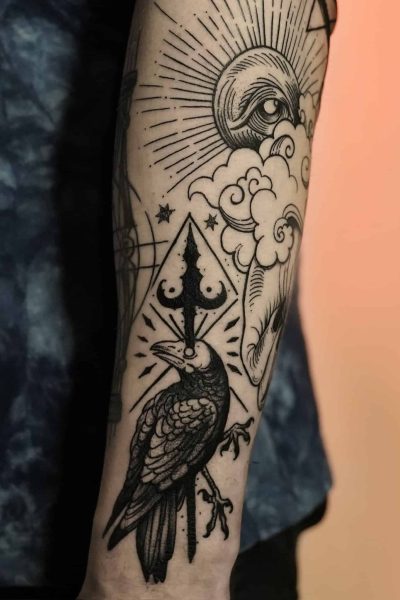 Zbliżenie tatuażu na przedramieniu z kolekcji DSC00032 7sinstattoo.live, przedstawiającego czarnego kruka siedzącego na gałęzi, z geometrycznym wzorem przypominającym włócznię nad głową. Wokół kruka znajdują się chmury, półksiężyc i skomplikowany wzór linii tworzący wzór oka i rozbłysku słońca. Osoba ma na sobie ciemnoniebieską koszulę.