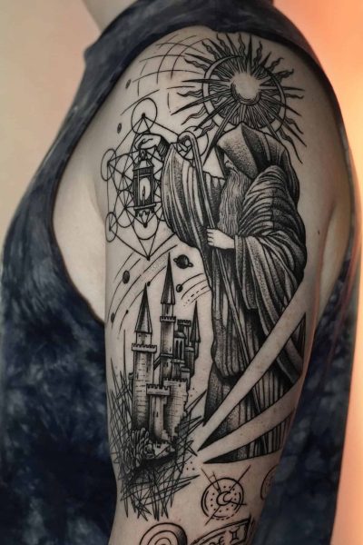 Na górnej części ramienia znajduje się szczegółowy czarno-szary tatuaż uchwycony jako DSC00054 przez 7sinstattoo.live. Wzór przedstawia postać w szacie trzymającą latarnię, stojącą nad zamkiem z trzema wieżami. Skomplikowane tło obejmuje geometryczne kształty, słońce z promieniami i różne abstrakcyjne elementy. Osoba ma na sobie ciemną koszulę bez rękawów. Aby zobaczyć podobne arcydzieła, odwiedź nasze wiodące studio tatuażu w Poznaniu, Seven Sins Tattoo.