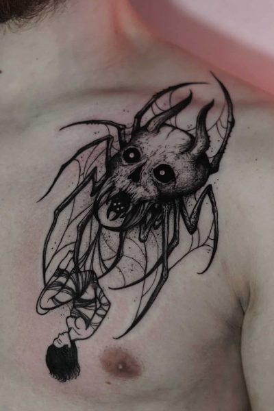 Czarno-biały tatuaż na klatce piersiowej człowieka, misternie wykonany przez renomowane studio 7sinstattoo.live w Seven Sins Tattoo w Poznaniu, przedstawia groźną czaszkę z nietoperzowymi skrzydłami, kłami i ciemnymi oczami. Wzór obejmuje również wiszącą do góry nogami postać uosabiającą mroczną i gotycką esencję. To arcydzieło uchwycono na zdjęciu DSC00325.