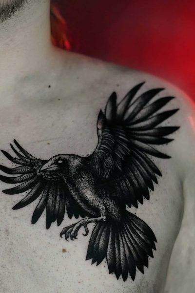 Szczegółowy i ciemny tatuaż kruka z szeroko rozpostartymi skrzydłami jest umieszczony w pobliżu lewego ramienia na górnej części klatki piersiowej mężczyzny. Tło jest mglistym, czerwonym gradientem, który podkreśla kunszt poznańskich profesjonalnych artystów tatuażu ze studia Seven Sins Tattoo.