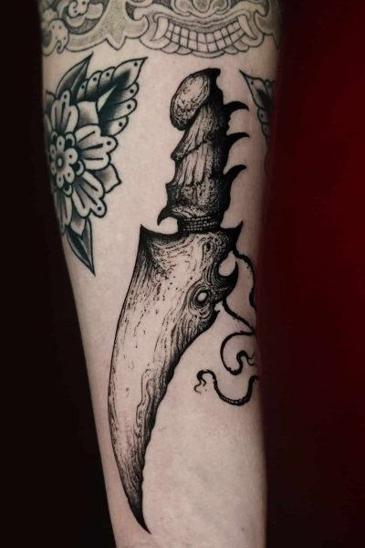 Czarno-biały tatuaż na przedramieniu przedstawia misternie zaprojektowany „7sinstattoo.live DSC00622 (1)” z Seven Sins Tattoo Studio w Poznaniu. Przedstawia zakrzywiony, ozdobny sztylet z rękojeścią ozdobioną szczegółowymi wzorami przypominającymi wielookie stworzenie z małymi mackami. Otaczające motywy kwiatowe wzmacniają ogólną kompozycję artystyczną stworzoną przez utalentowanego tatuażystę z Poznania.