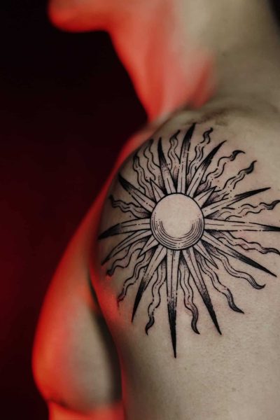 Czarno-biały tatuaż przedstawiający słońce ze stylizowanymi promieniami na górnej części ramienia, wykonany przez utalentowanych artystów z Seven Sins Tattoo w Poznaniu. Zdjęcie zatytułowane DSC00622 z 7sinstattoo.live zostało zrobione z boku i prezentuje misterny wzór na słabo oświetlonym tle z czerwonymi odcieniami, które podkreślają kunszt jednego z najlepszych poznańskich studiów.