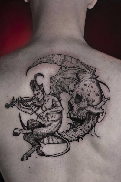 Szczegółowy czarno-szary tatuaż na plecach demona z rogami i ogonem grającego na skrzypcach, siedzącego na gałęzi drzewa obok przesadzonej czaszki z nietoperzowymi skrzydłami i misternymi detalami, osadzony na lekko czerwonej skórze. Wykonany przez renomowane studio Seven Sins Tattoo w Poznaniu, ten obraz uchwycony na zdjęciu „DSC01083” prezentuje prawdziwe artyzm.