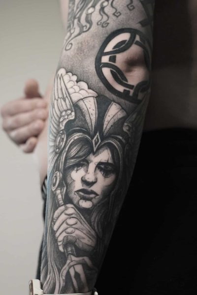 7sinstattoo.live Szczegółowy czarno-szary tatuaż ze studia tatuażu Poznań, zidentyfikowany jako DSC03406, pokrywa przedramię osoby. Przedstawia zaciekłą, wojowniczkę w hełmie ze skrzydłami na hełmie i skupionym wyrazem twarzy, trzymającą miecz. Dodatkowe abstrakcyjne elementy są widoczne wokół tatuażu, rozciągające się w górę ramienia. Najlepsze studio tatuażu Seven Sins Tattoo