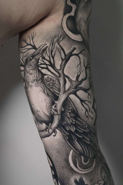 7sinstattoo.live Szczegółowy czarno-szary tatuaż, oznaczony jako DSC03442 i stworzony w Najlepszym Studio Tatuażu Poznań, przedstawia kruka siedzącego na sękatej gałęzi drzewa na ramieniu człowieka. Wzór charakteryzuje się misternym cieniowaniem i cienkimi liniami, dzięki czemu zarówno ptak, jak i gałąź wyglądają realistycznie. Najlepsze studio tatuażu Seven Sins Tattoo