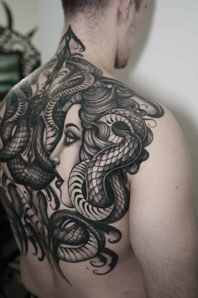 7sinstattoo.live Osoba z dużym, szczegółowym tatuażem na plecach, przedstawiającym twarz kobiety przeplataną kilkoma realistycznie wyglądającymi wężami, wybrała DSC03480 ze względu na skomplikowane cieniowanie i faktury. Tatuaż rozciąga się od ramienia do ramienia i w dół do dolnej części pleców, prezentując kunszt najlepszego studia tatuażu Poznań. Najlepsze studio tatuażu Seven Sins Tattoo