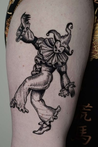 7sinstattoo.live Zbliżenie na ramię osoby pokazuje szczegółowy czarno-szary tatuaż błazna w klasycznym stroju z falbaniastym kołnierzem, kapeluszem i spiczastymi butami. Błazen jest przedstawiony w animowanej, żywej pozie, emanującej kaprysem. Uchwycony jako DSC04234 w tatuaż Poznań, osoba ma na sobie ciemne, wzorzyste ubranie. Najlepsze studio tatuażu Seven Sins Tattoo