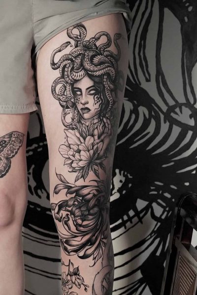 7sinstattoo.live Wytatuowana noga z dużą głową Meduzy z włosem węża, różą i innymi skomplikowanymi wzorami, takimi jak ćma i krab, rozciąga się od połowy uda do kolana. Szczegółowe monochromatyczne dzieło sztuki wyróżnia się na tle czarnego muralu, wszystko fachowo wykonane w studiu DSC04304-восстановлено w Poznaniu. Najlepsze studio tatuażu Seven Sins Tattoo