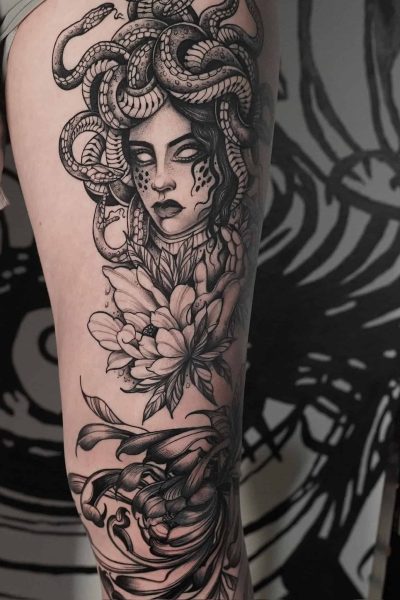 7sinstattoo.live Czarno-biały tatuaż na górnej części uda osoby przedstawiający głowę Meduzy z włosami węża, otoczony kwiatowymi wzorami i skomplikowanymi wzorami. Szczegółowe dzieło sztuki, wykonane w najlepszym studio tatuażu w Poznaniu, obejmuje znaczną część nogi. | Nazwa produktu: DSC04312 Najlepsze studio tatuażu Seven Sins Tattoo