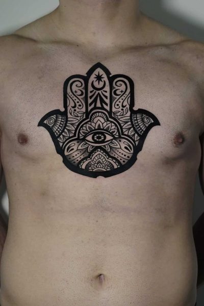 7sinstattoo.live Osoba z dużym, misternym czarnym tatuażem symbolu Hamsa na piersi stoi na prostym tle. Wzór charakteryzuje się ozdobnymi wzorami i obejmuje oko w środku dłoni, rozciągające się na górną część klatki piersiowej. Ta oszałamiająca praca została uchwycona w produkcie DSC04471 w studio tatuażu Poznań. Najlepsze studio tatuażu Seven Sins Tattoo