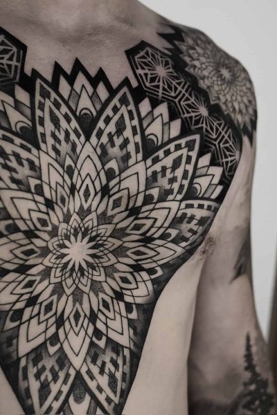 7sinstattoo.live Zbliżenie górnej części klatki piersiowej i ramion osoby prezentuje rozległy i skomplikowany geometryczny wzór tatuażu z najlepszego studia tatuażu Poznań, zidentyfikowanego po nazwie produktu DSC04832. Tatuaż charakteryzuje się symetrycznymi wzorami i kształtami przypominającymi mandalę z czarnym i szarym cieniowaniem, pokrywającymi znaczną część skóry. Najlepsze studio tatuażu Seven Sins Tattoo
